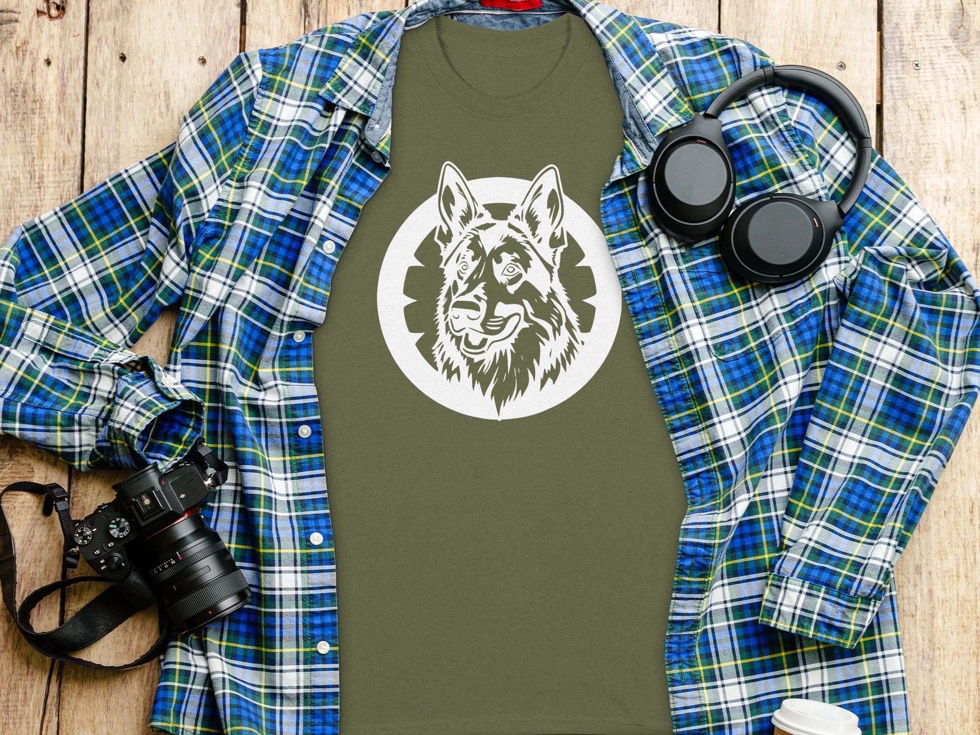 Green t-shirt with a white German Shepherd graphic, laid out with a plaid shirt, headphones, and camera.
