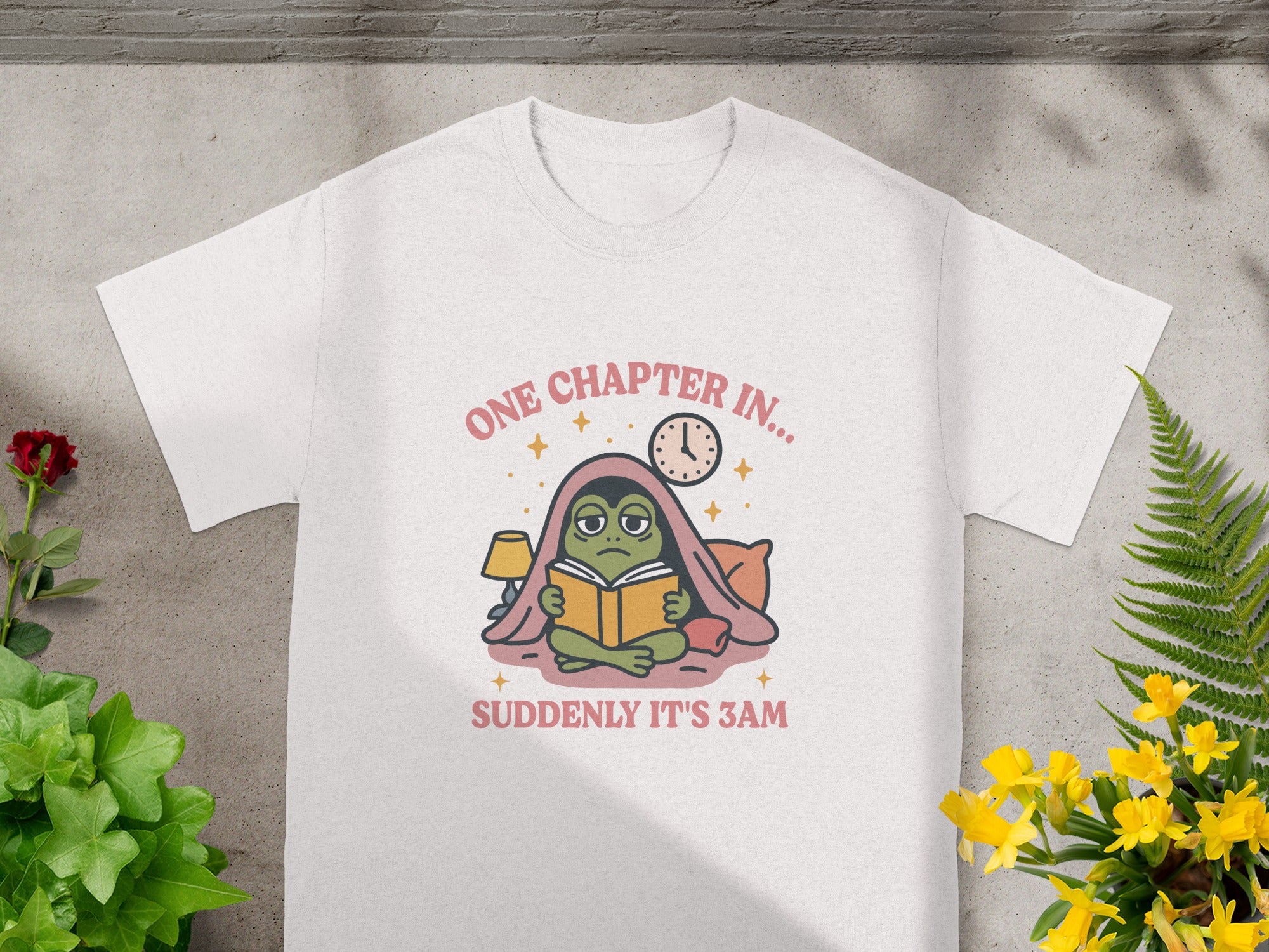 White t-shirt with illustration of a frog reading a book, "ONE CHAPTER IN...", and "SUDDENLY IT'S 3AM".