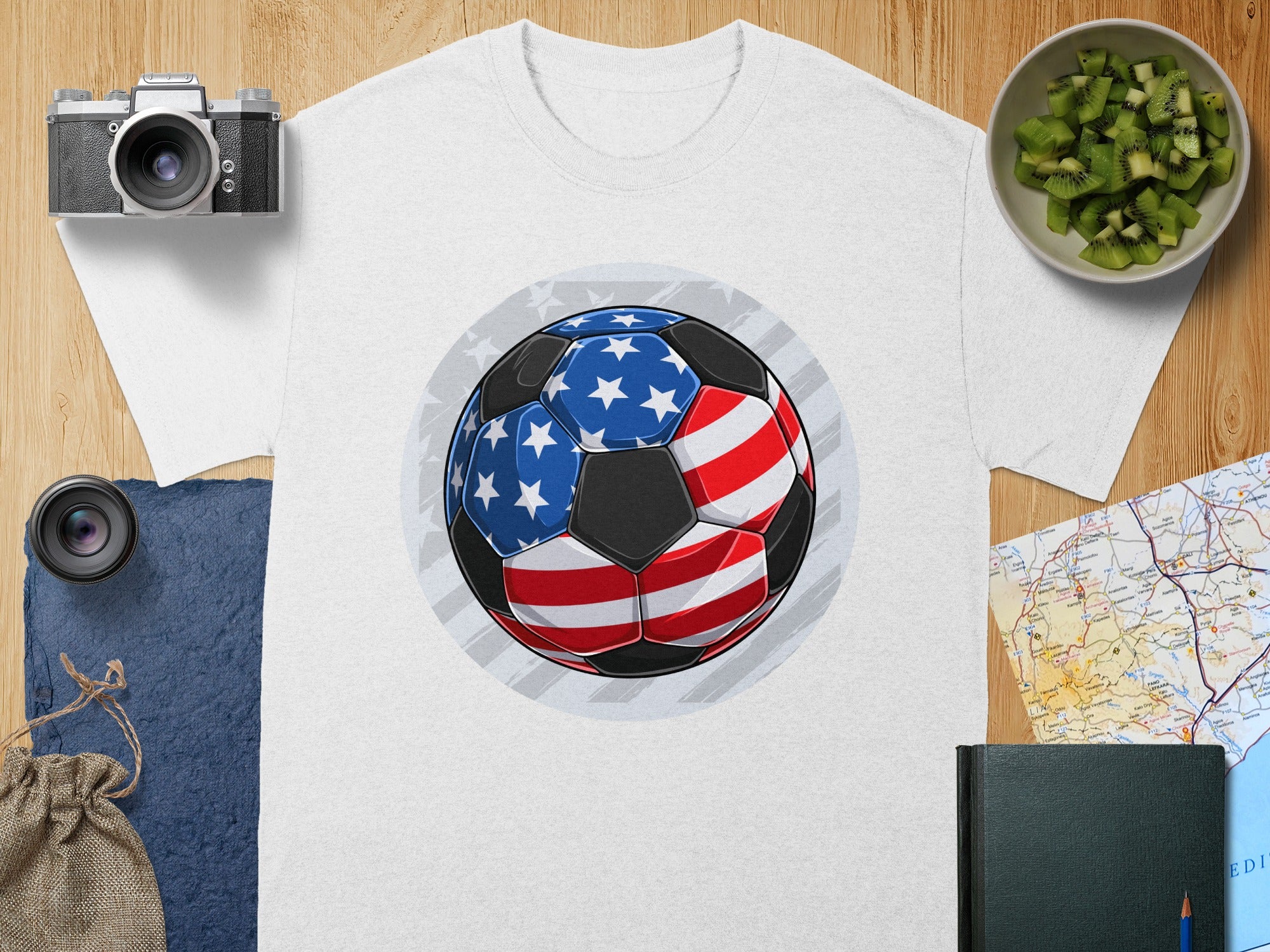 White t-shirt with an American flag soccer ball graphic on a wooden surface.
