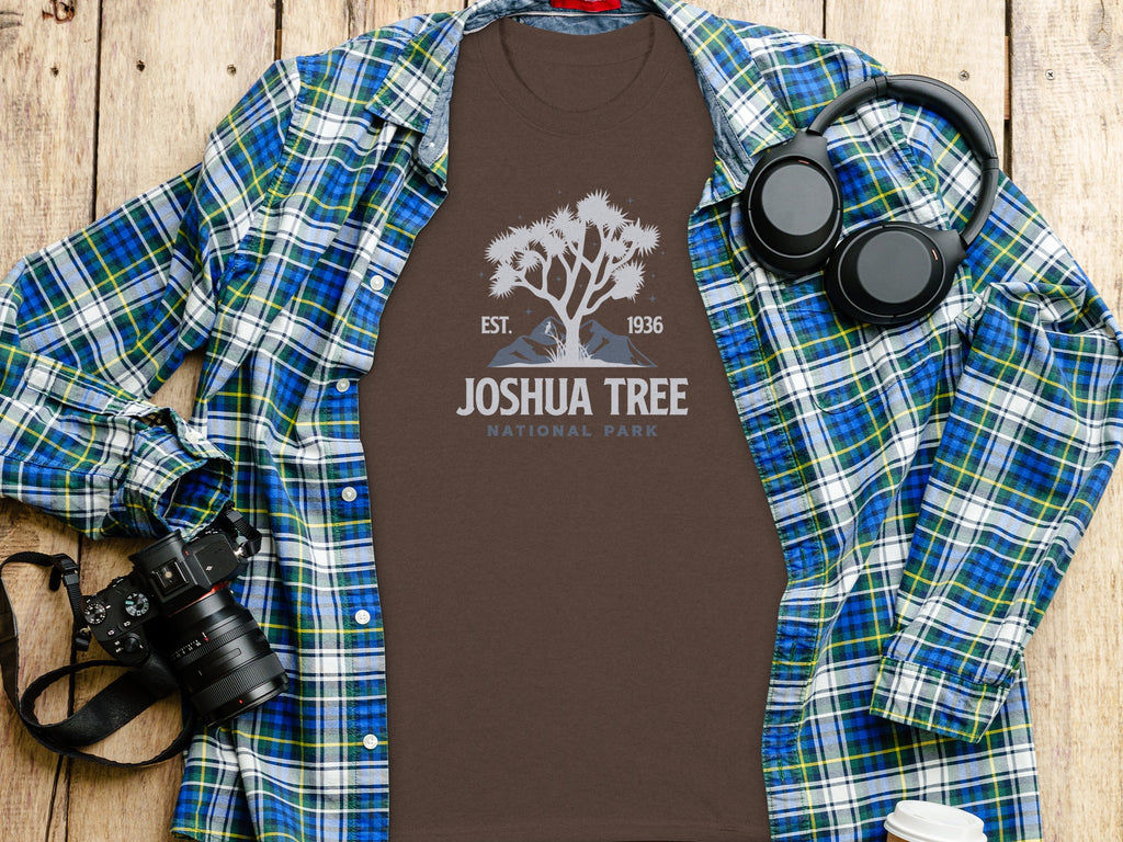 Brown t-shirt with Joshua Tree National Park graphic, laid on wooden surface with flannel shirt and headphones.