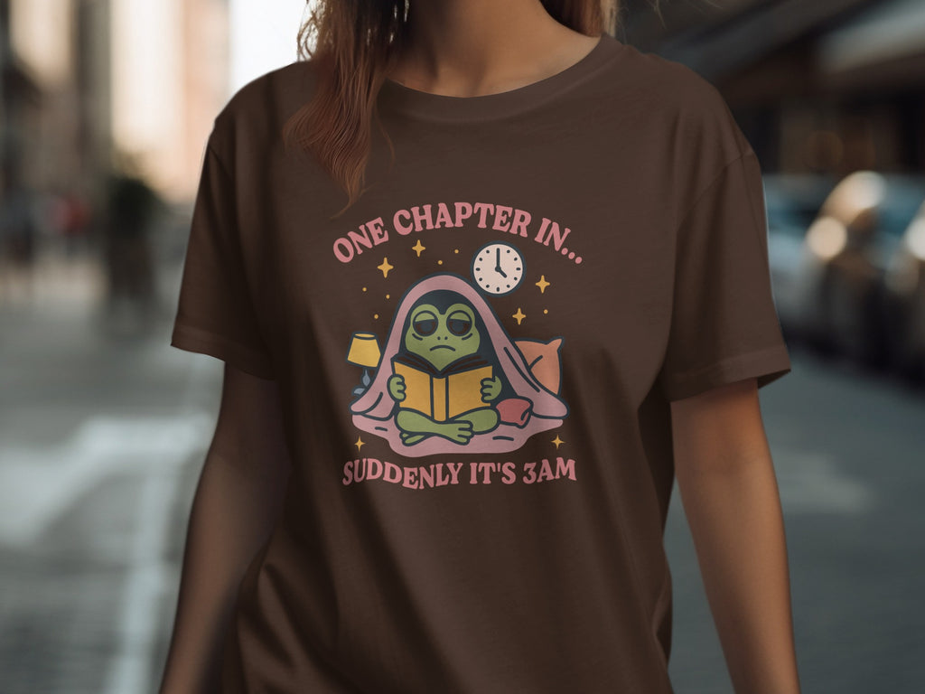 Brown t-shirt with frog reading under blankets, "One chapter in... suddenly it's 3am."