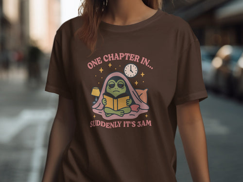 Brown t-shirt with frog reading under blankets, "One chapter in... suddenly it's 3am."