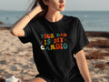 Black t-shirt with retro text "YOUR DAD IS MY CARDIO" on a beach.