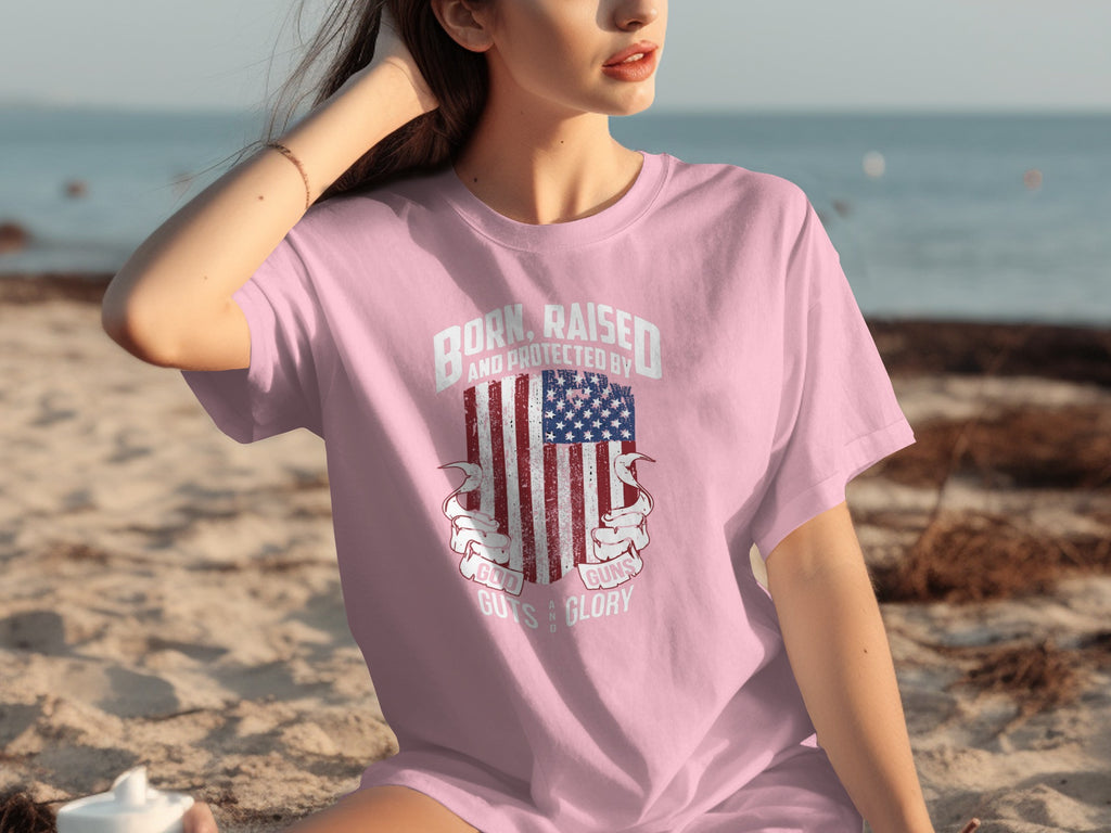 Pink t-shirt with American flag graphic and text: "Born, raised, and protected by God, guns, guts, and glory."
