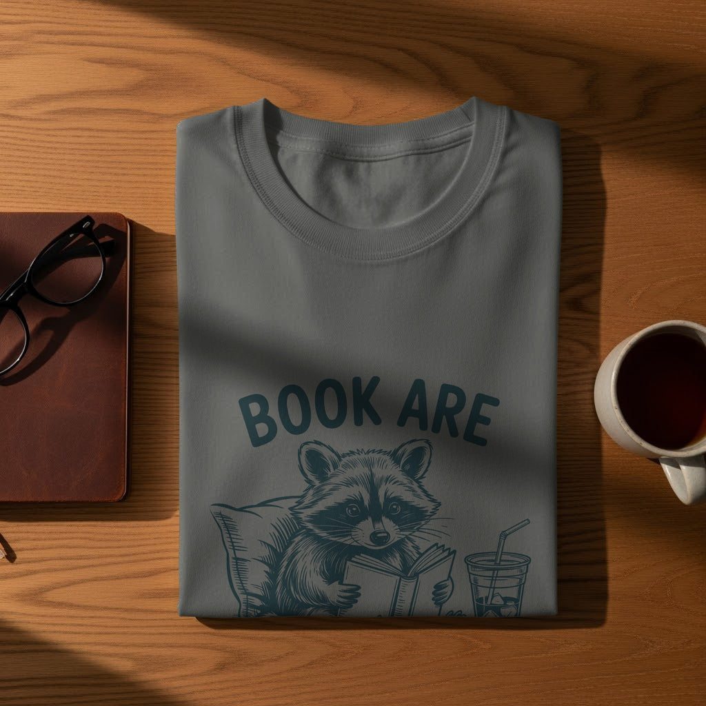 Gray t-shirt with raccoon reading a book and drinking an iced beverage.