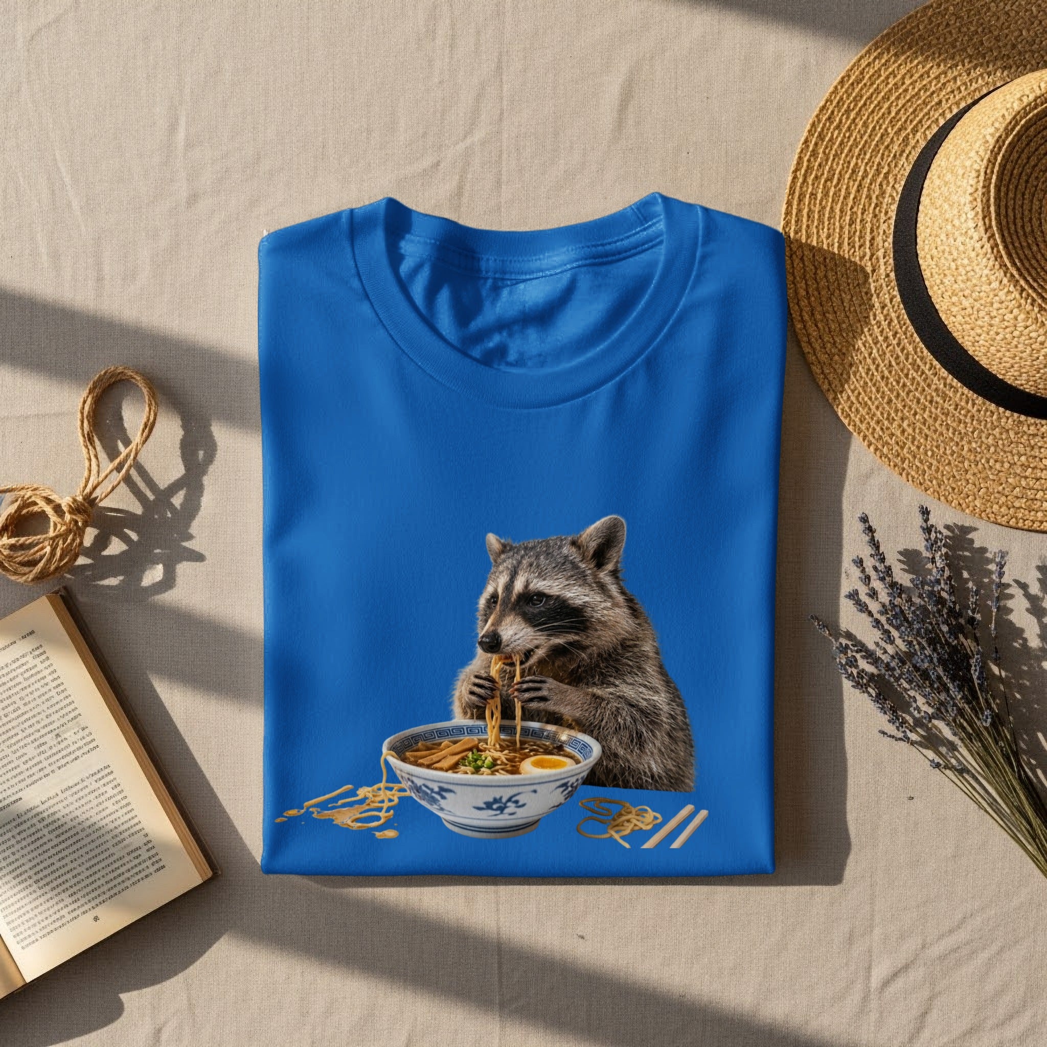 Blue t-shirt featuring a raccoon eating ramen noodles from a bowl.