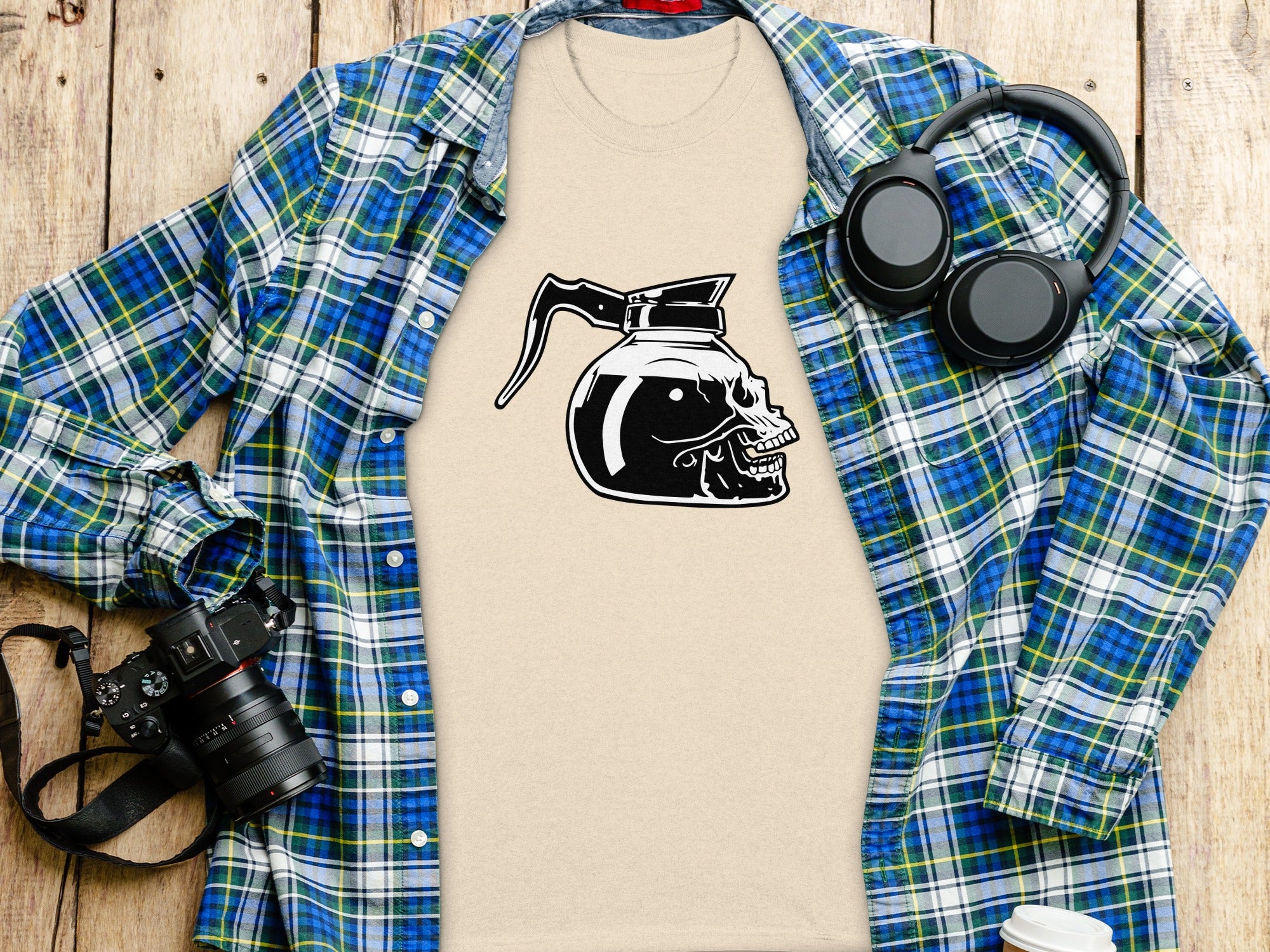 Beige t-shirt with a black skull and coffee pot graphic, laid out with a plaid shirt.