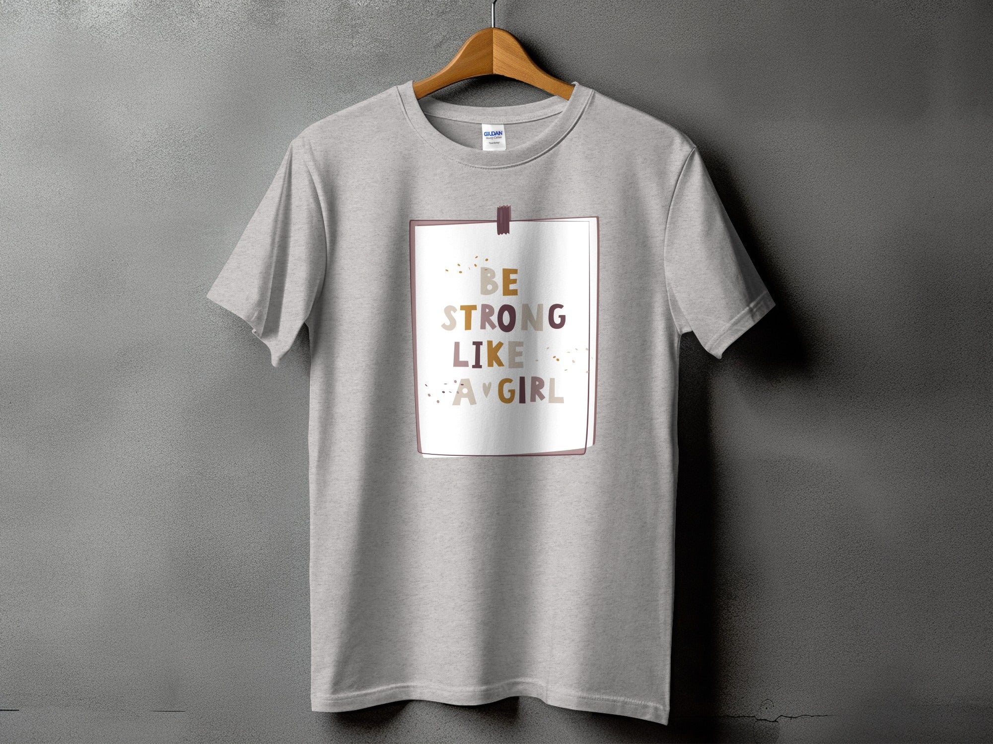 Grey t-shirt with "Be Strong Like A Girl" text in muted colors inside a framed border.