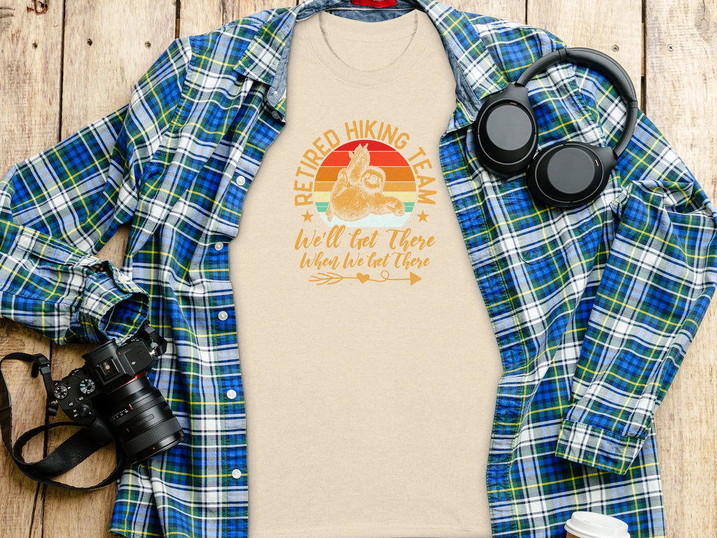 Tshirt with "Retired Hiking Team" printed on it, next to a plaid shirt, headphones, and camera.