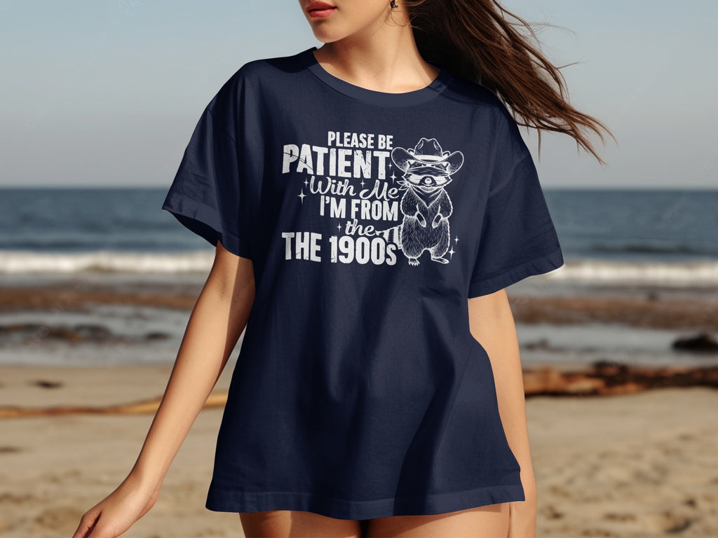 Navy t-shirt with white text and drawing of a raccoon in cowboy hat saying "Please be patient with me I'm from the 1900s".