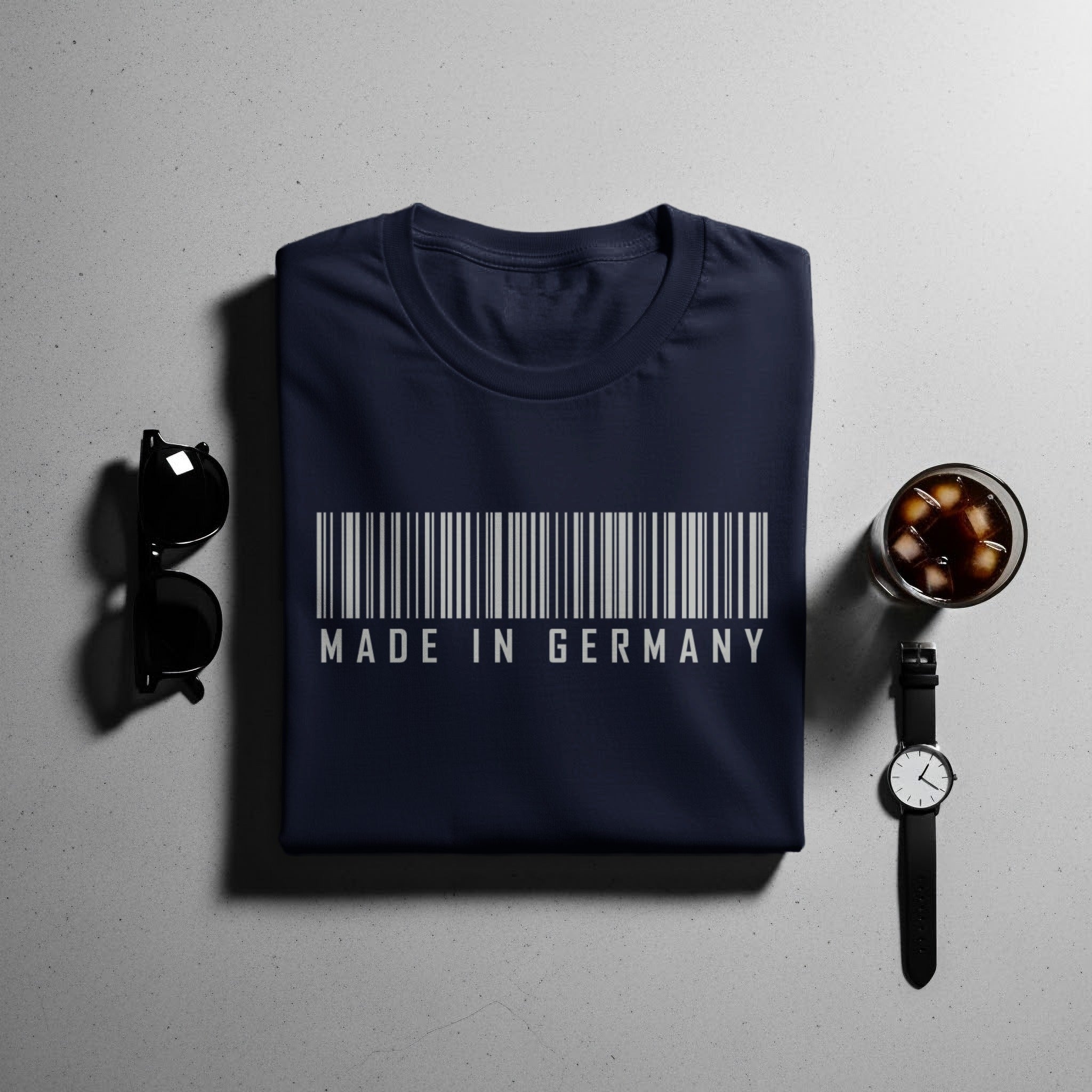 Navy t-shirt with barcode and "Made in Germany" text, with sunglasses, drink, and watch.