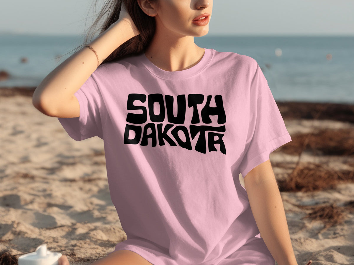 Pink t-shirt with black "SOUTH DAKOTA" text, worn by a woman on a beach.