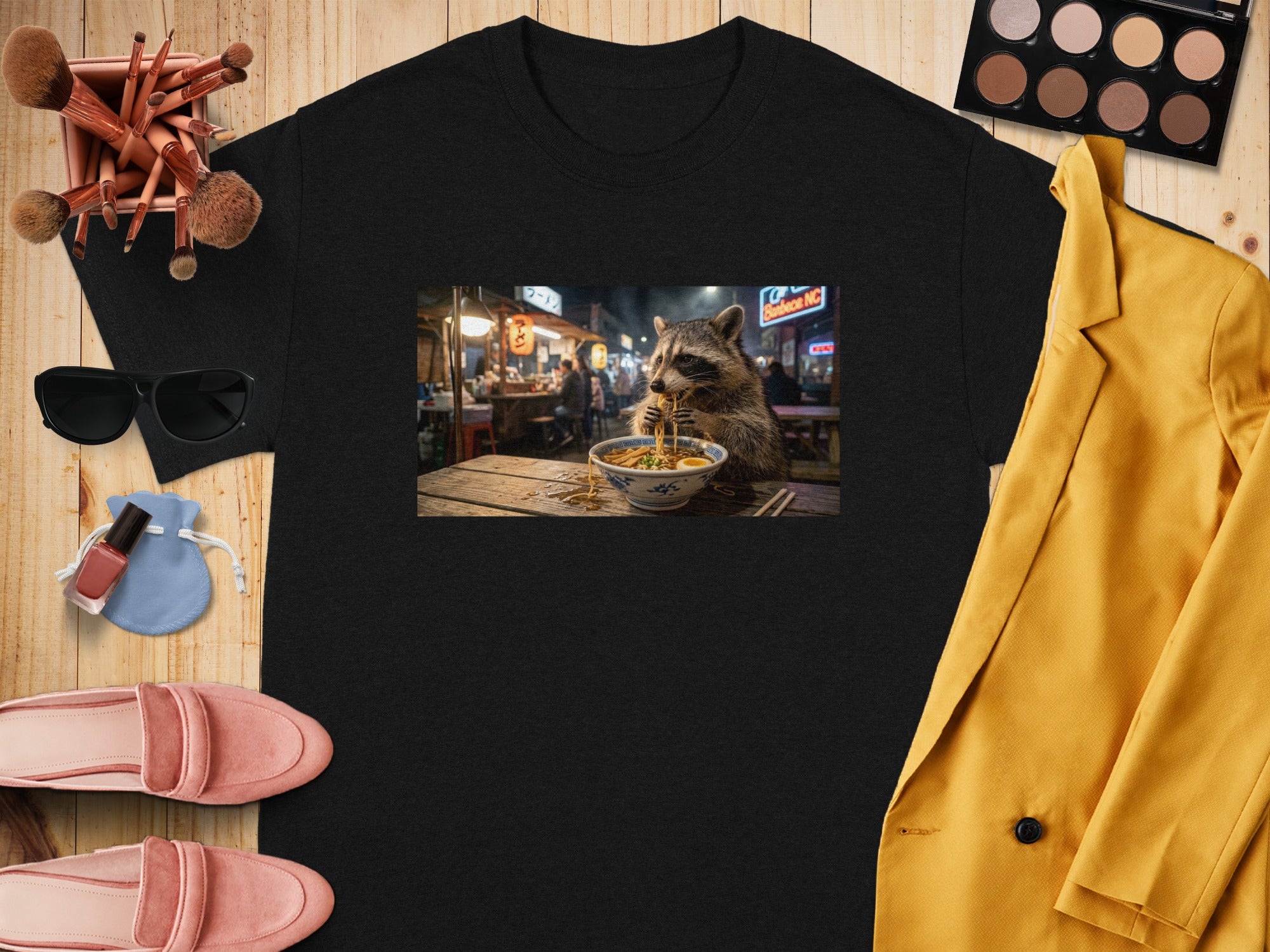Black t-shirt with a graphic of a raccoon eating ramen in a busy street market.