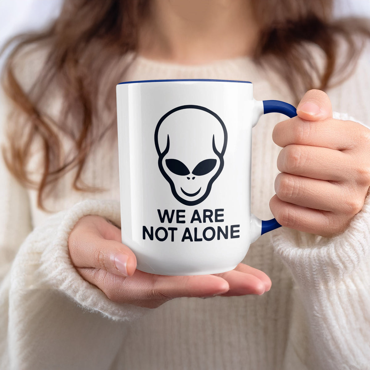 White mug with alien face and text "WE ARE NOT ALONE" held by hands.