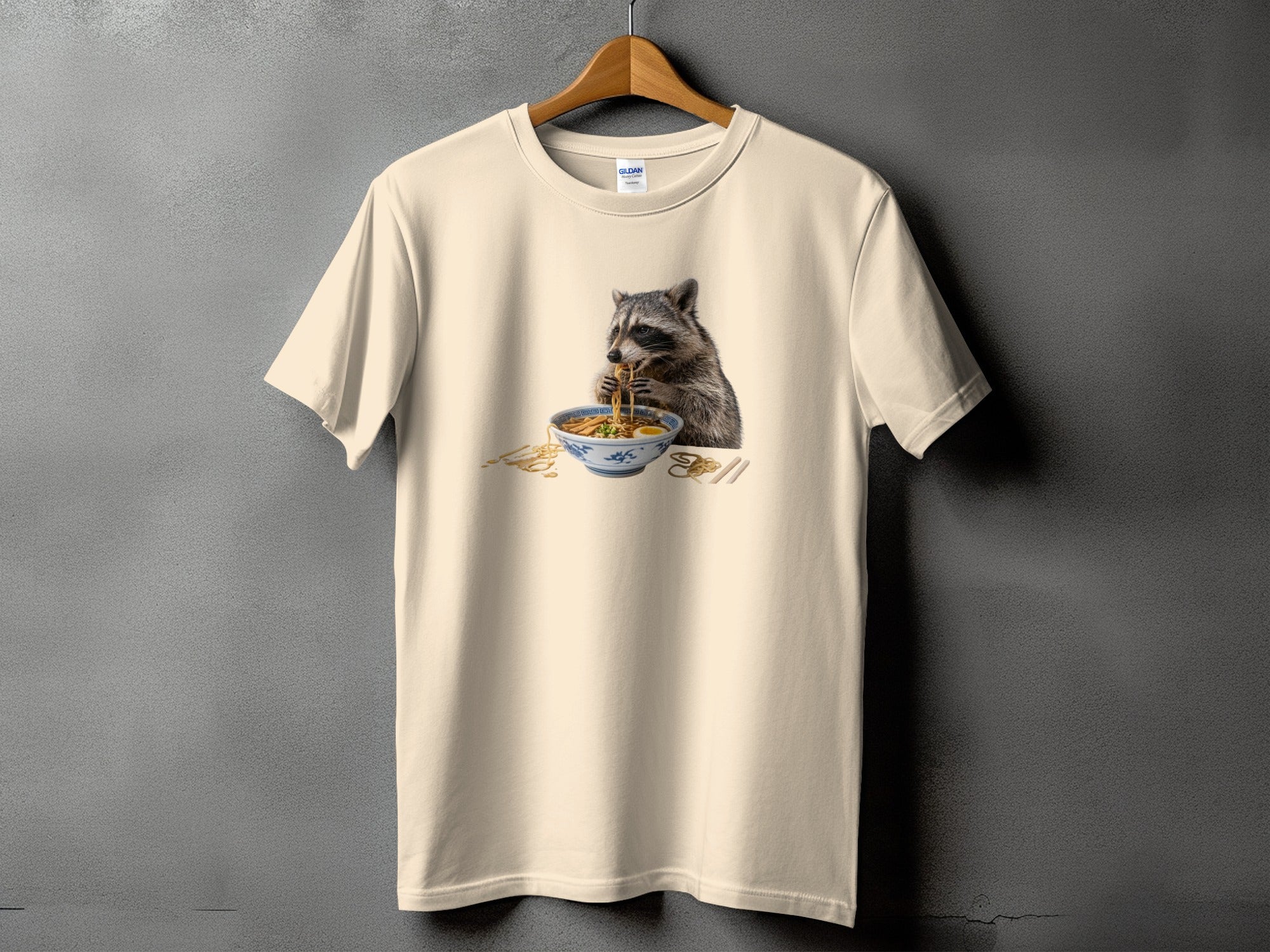 Cream t-shirt with a graphic of a raccoon eating ramen noodles from a bowl.