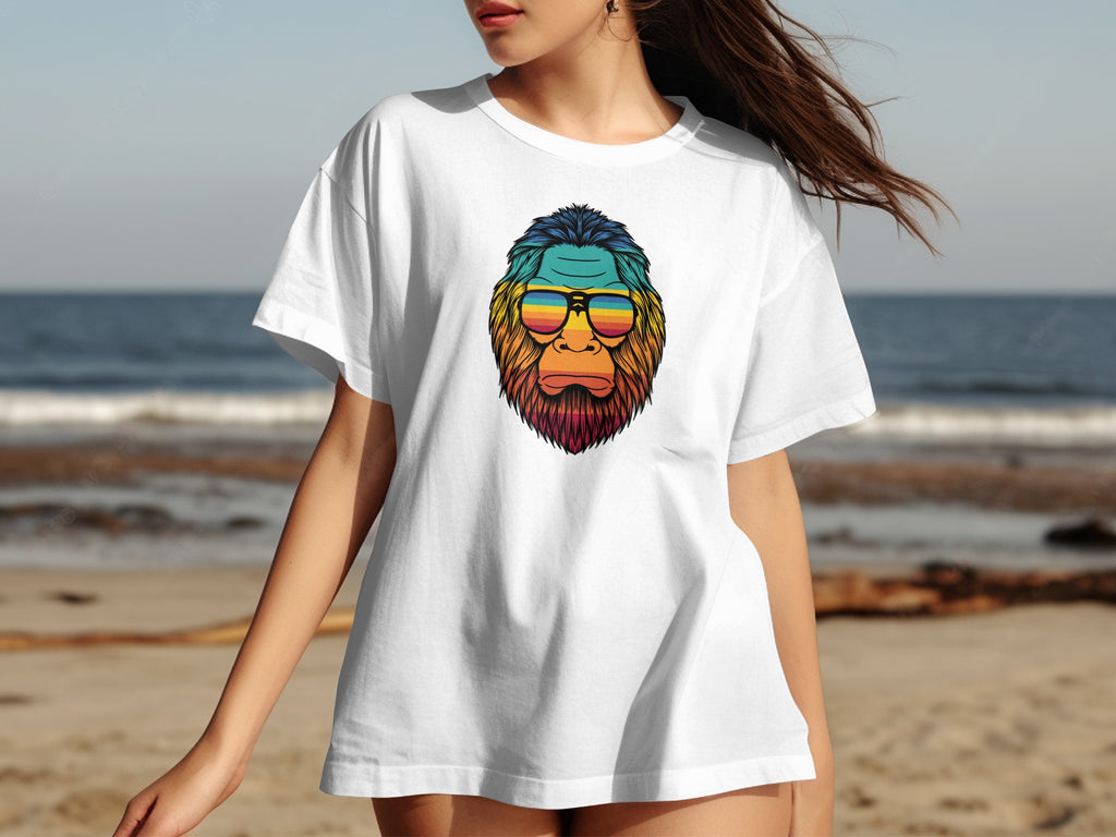 White t-shirt with a colorful Bigfoot graphic wearing sunglasses on a beach