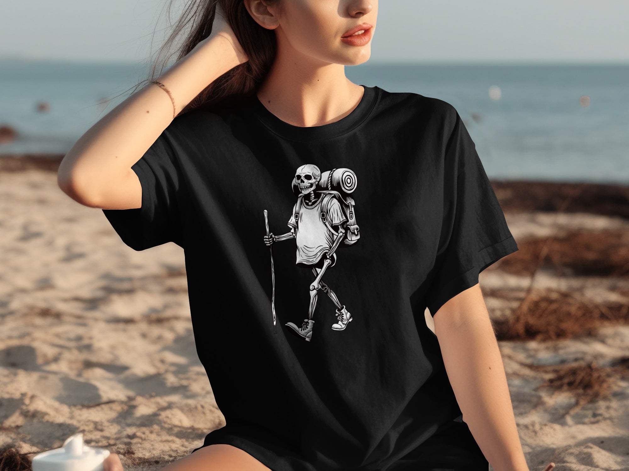 Black t-shirt with a skeleton hiker graphic, set against a beach background.