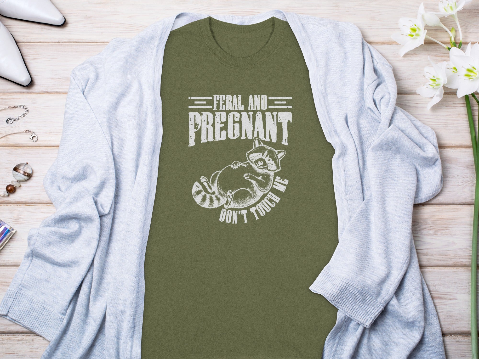 Olive t-shirt with white text "FERAL AND PREGNANT DON'T TOUCH ME" and raccoon graphic.