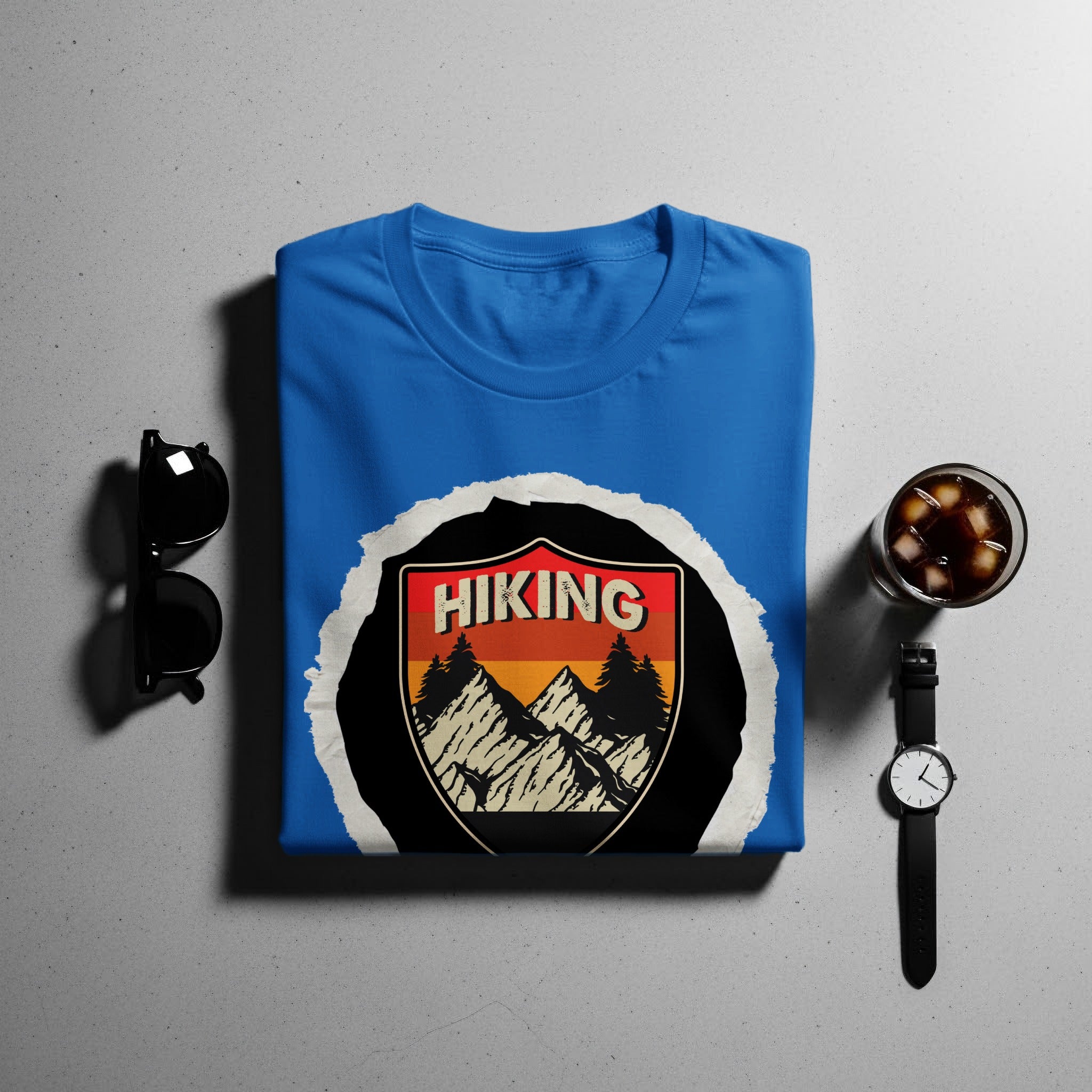 Blue t-shirt with "Hiking" mountain badge design, displayed with sunglasses and watch.