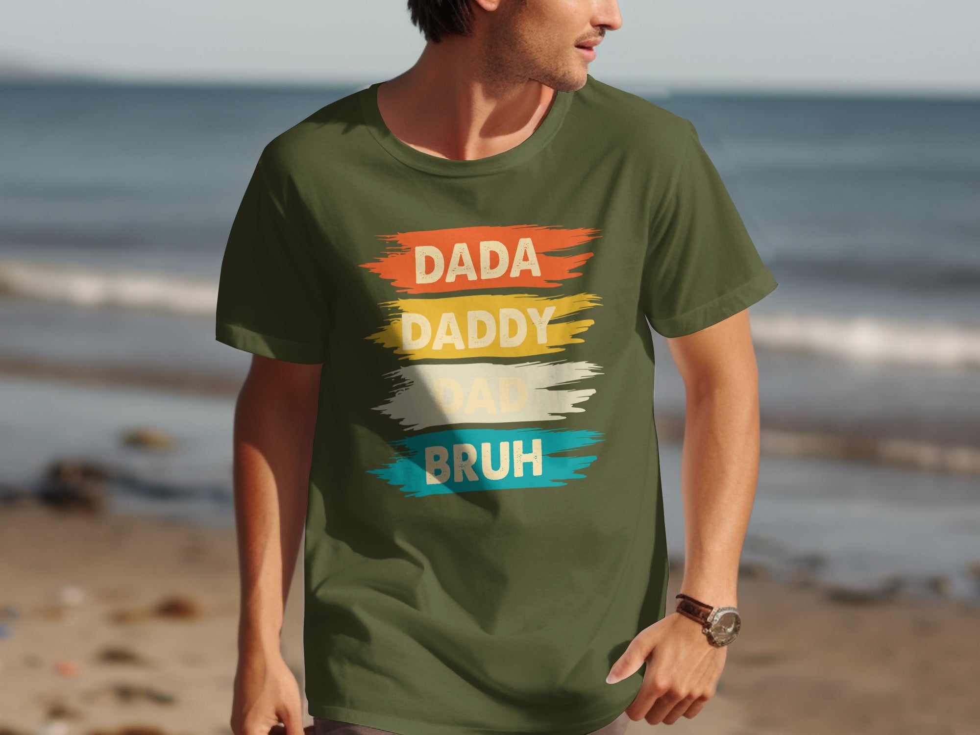 Olive green t-shirt with text "DADA DADDY DAD BRUH" in distressed, colorful brush strokes.