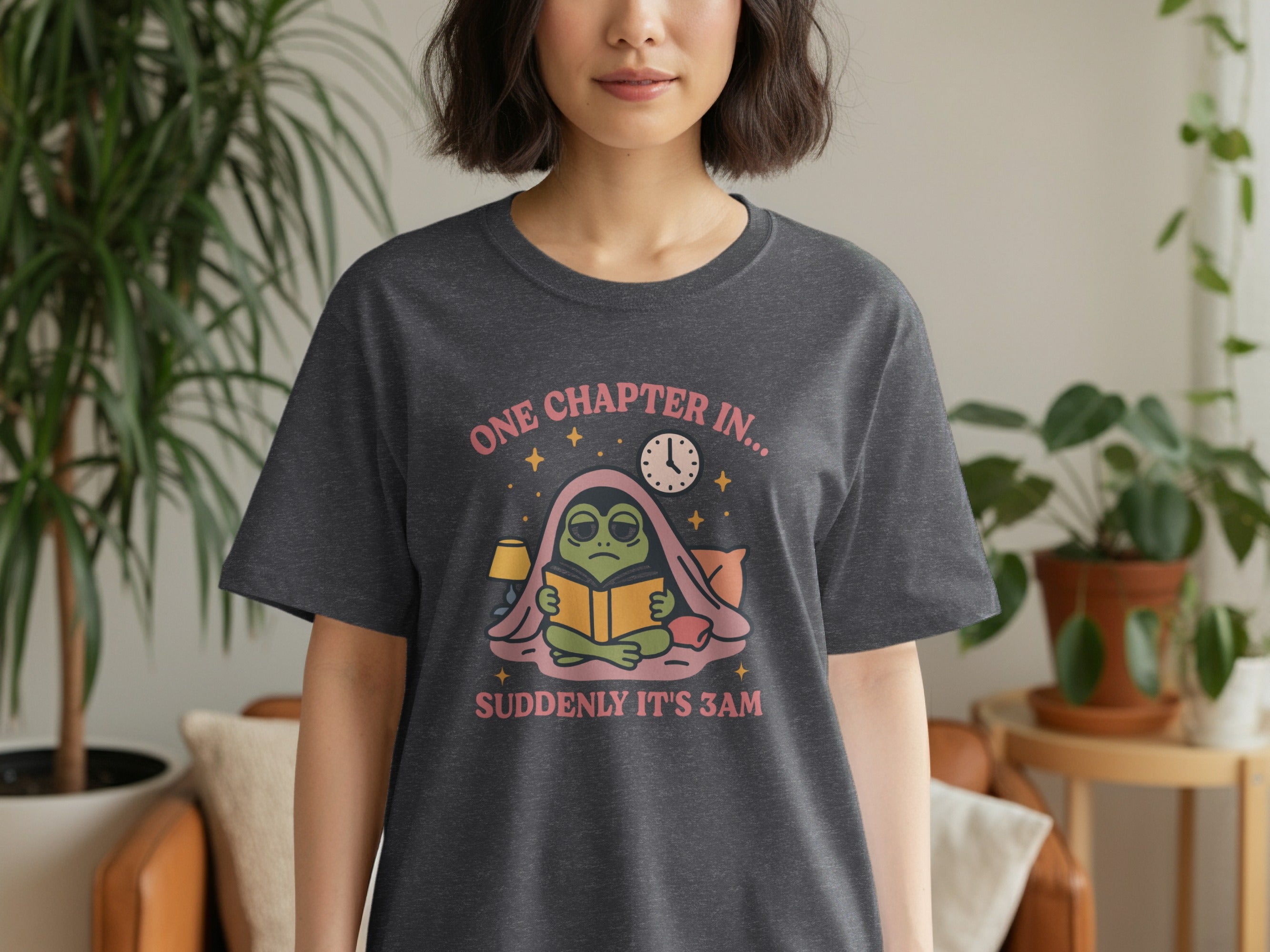 Graphic t-shirt featuring a frog reading a book under a blanket with text "ONE CHAPTER IN..." and "SUDDENLY IT'S 3AM".
