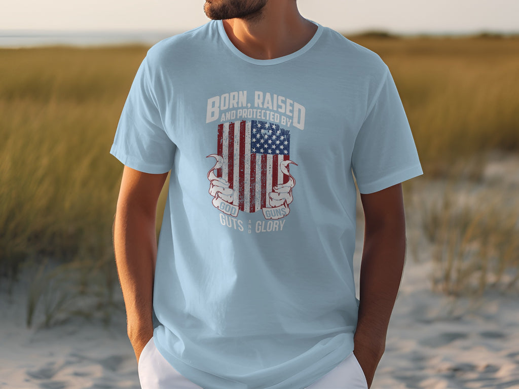Light blue t-shirt with American flag graphic and text reading "Born, Raised And Protected By God, Guns, Guts & Glory".