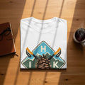White t-shirt with a nature graphic featuring a pinecone, mountains, ski poles, and letters H I K.