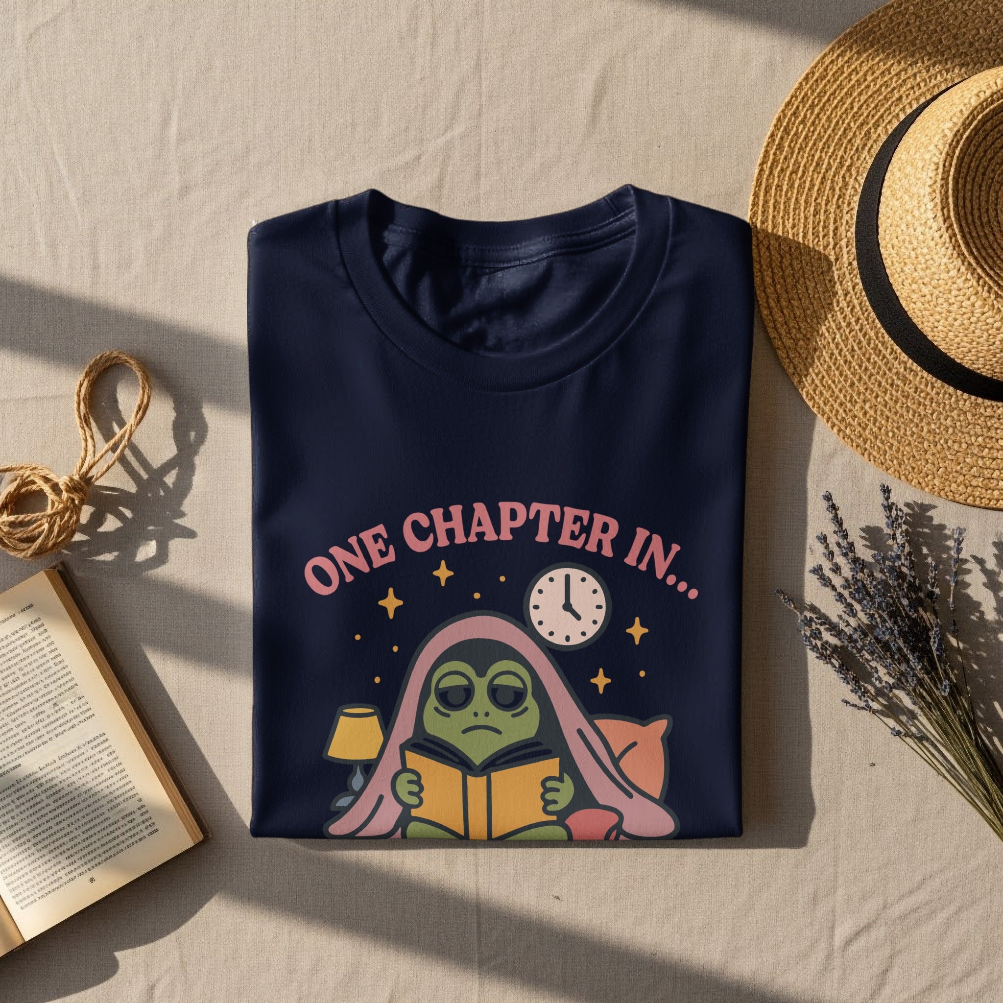 Navy t-shirt with a sleepy frog reading under the text "ONE CHAPTER IN...".