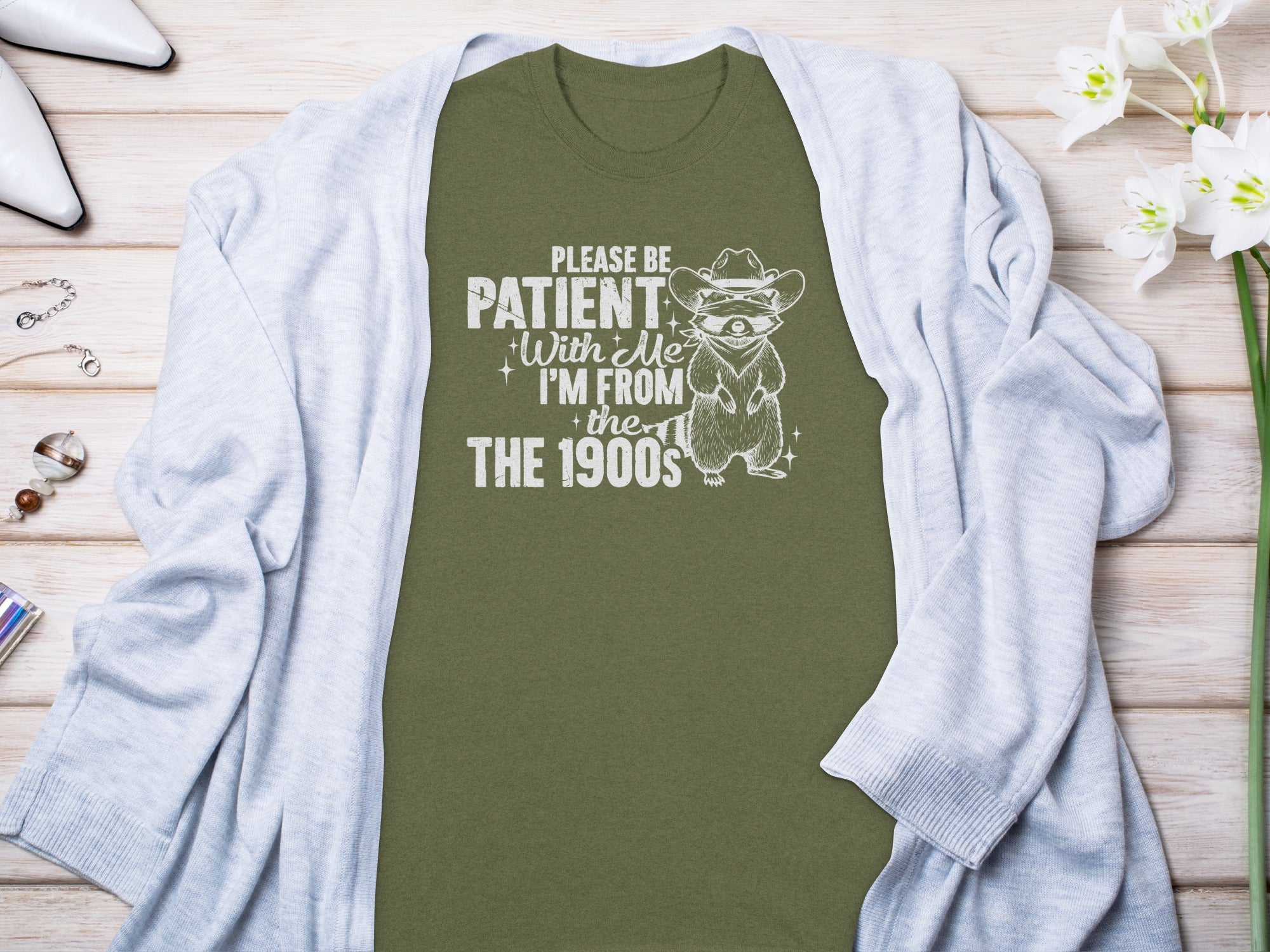 Olive green t-shirt with white text and raccoon graphic reading "PLEASE BE PATIENT With Me I'M FROM the THE 1900s".