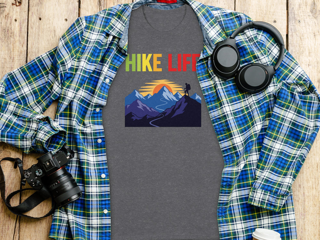 Grey t-shirt with "HIKE LIFE" text and mountain graphic, laid on a wooden surface with a plaid shirt, camera, and headphones.