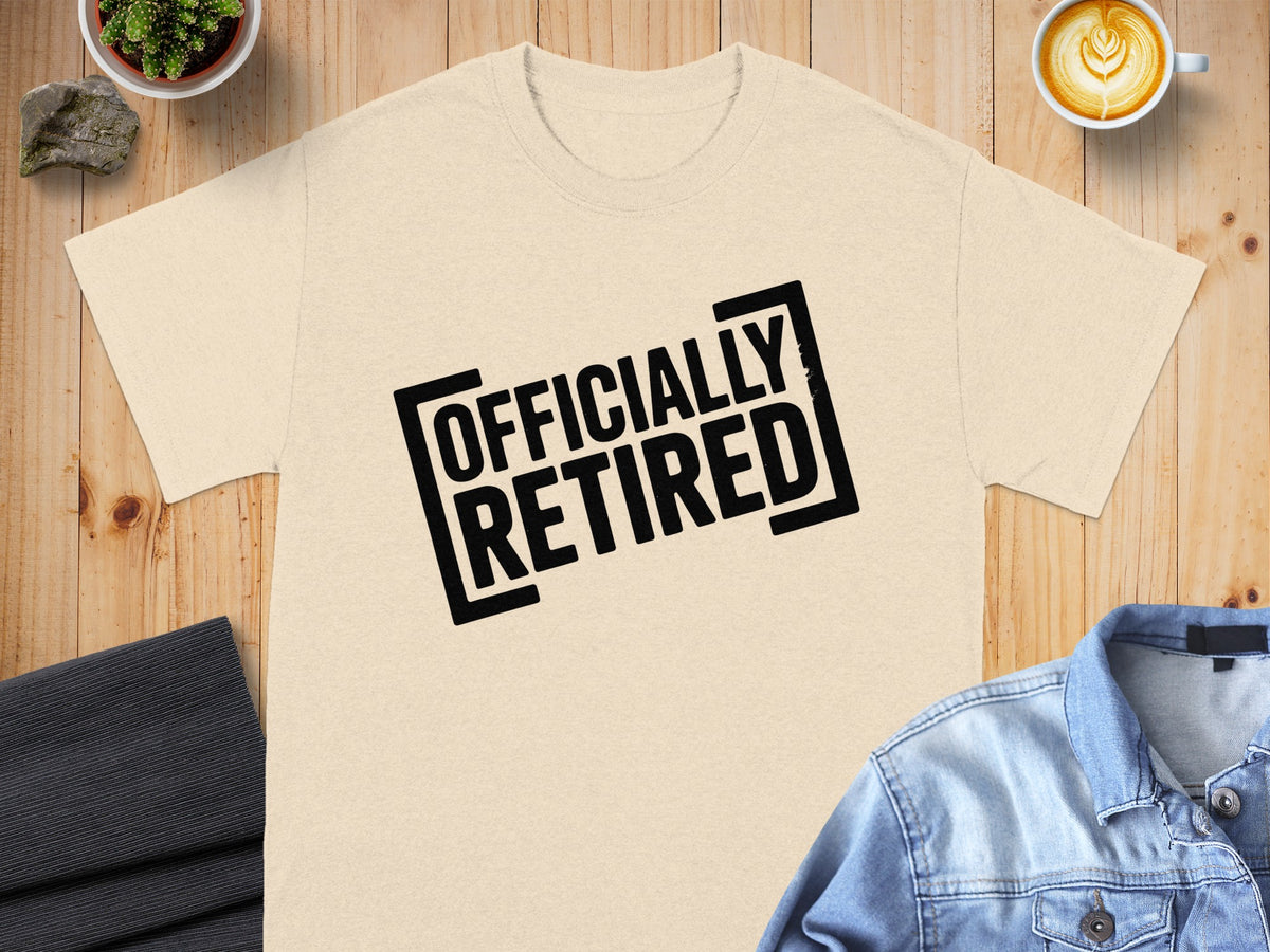 Cream t-shirt with black text "OFFICIALLY RETIRED" displayed on a wooden surface.