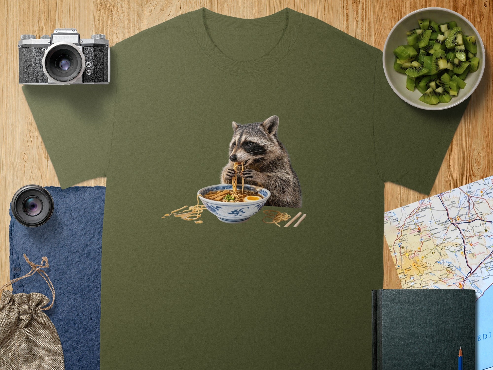 An olive-green t-shirt features a graphic of a raccoon eating a bowl of ramen noodles.
