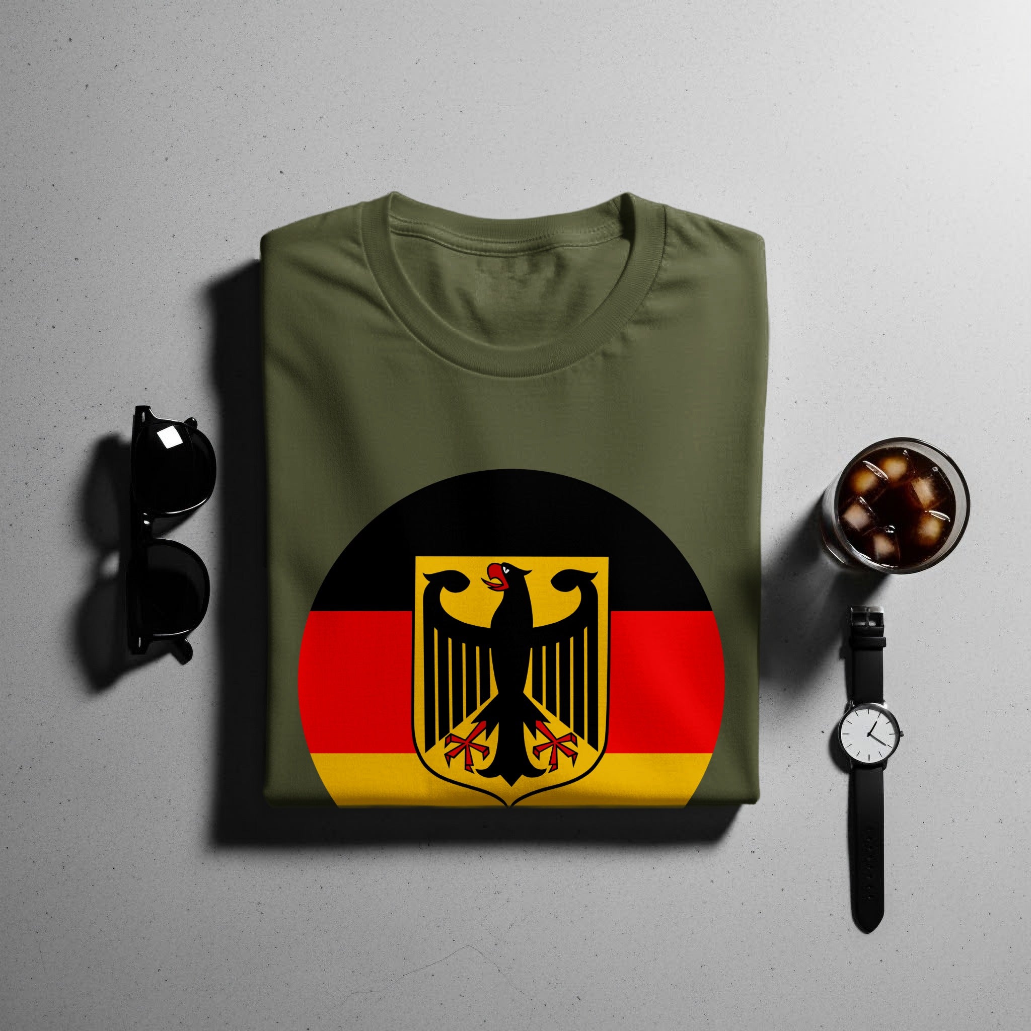 Olive green t-shirt with German flag emblem and black shield on a concrete background.