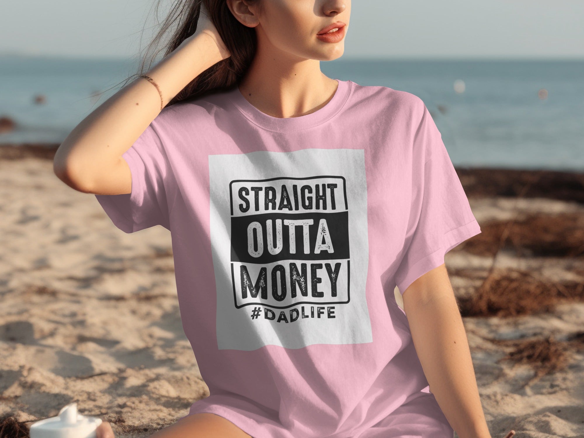 Pink t-shirt with "STRAIGHT OUTTA MONEY #DADLIFE" graphic on a beach.