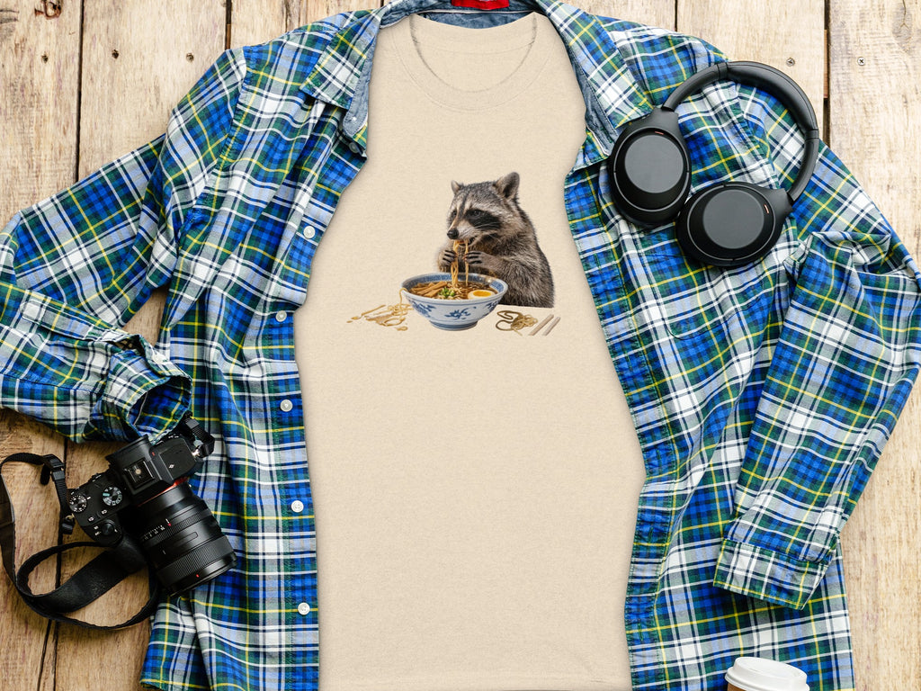 T-shirt featuring a raccoon eating ramen, laid flat with a flannel shirt.