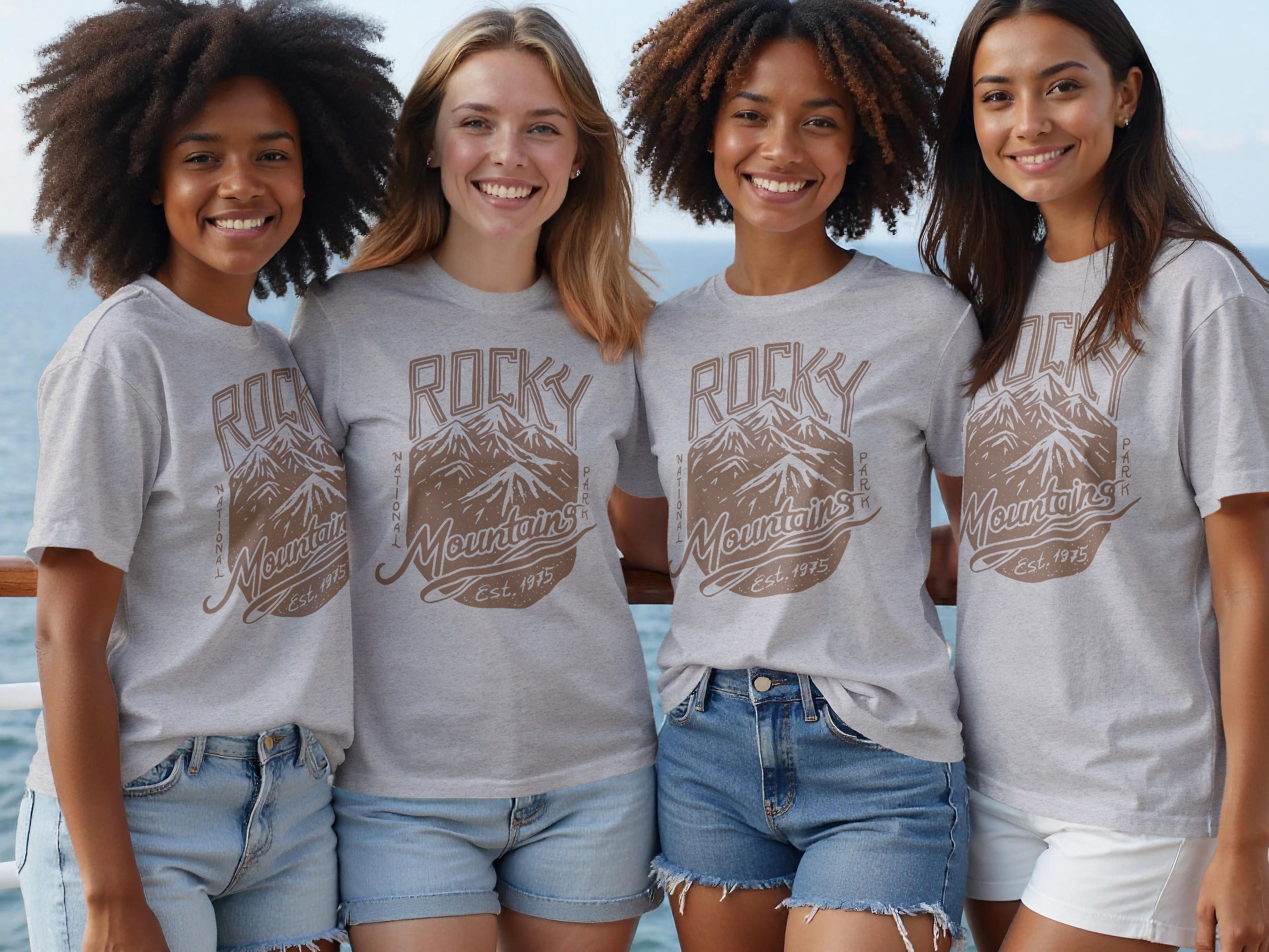 Four smiling women wear grey t-shirts with a "Rocky Mountains National Park Est. 1975" mountain graphic.