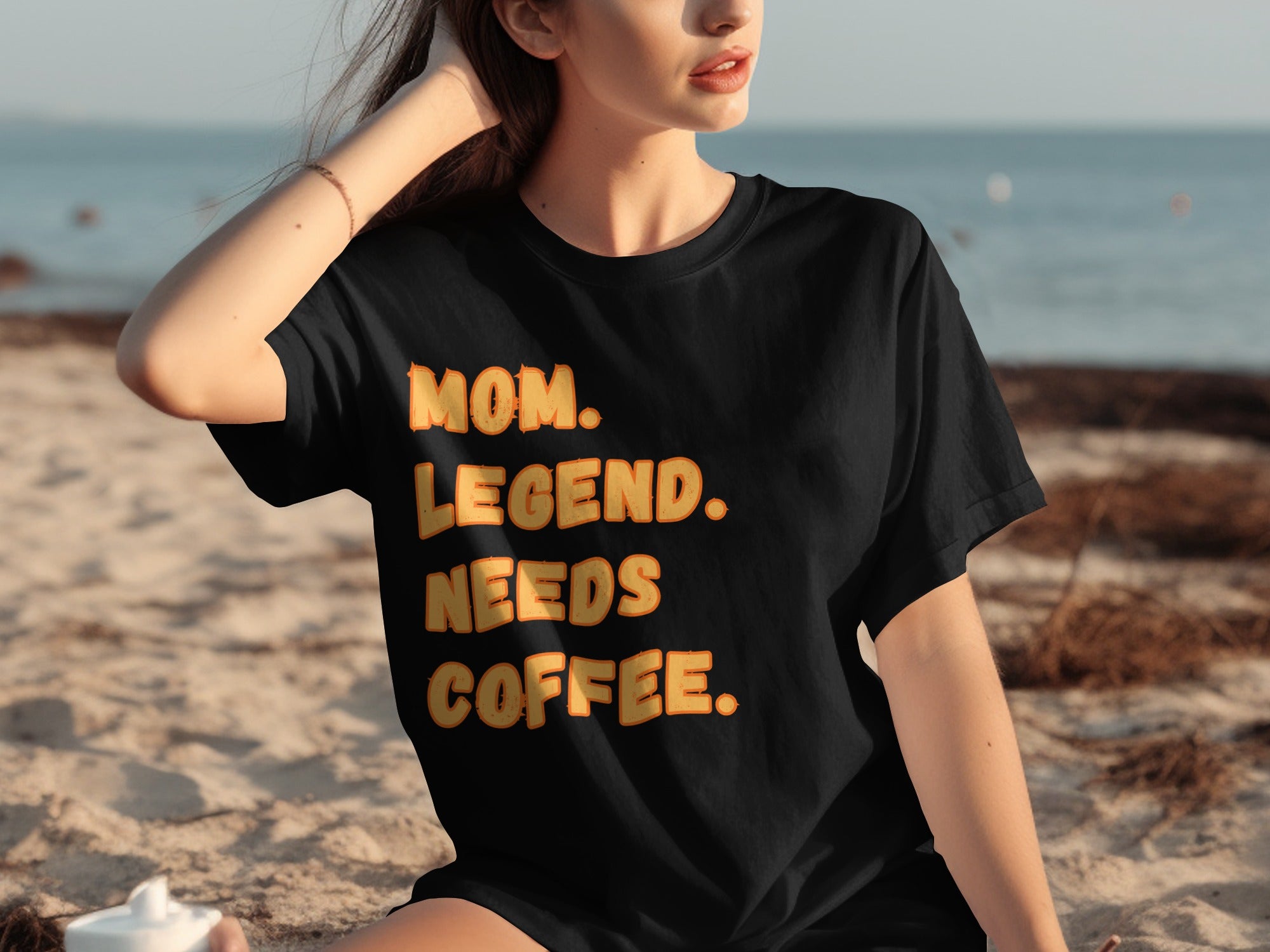 Black t-shirt with text "MOM. LEGEND. NEEDS COFFEE." on a beach.